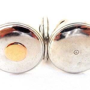 May include: A silver-toned antique pocket watch with a gold-colored circular accent on the front. The watch has a hinged design, with two circular faces connected by a small hinge. The watch face has a textured, circular pattern.