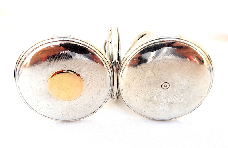 May include: A silver-toned antique pocket watch with a gold-colored circular accent on the front. The watch has a hinged design, with two circular faces connected by a small hinge. The watch face has a textured, circular pattern.