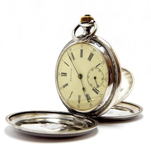 May include: A silver Zenith pocket watch with an open face and a cream-colored dial. The watch features black Roman numerals, a small seconds dial, and a gold-colored crown. The watch's cover is open, revealing the inner workings.