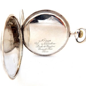May include: Open silver pocket watch with the back cover ajar, revealing the watch face. The watch has a silver case and a gold-colored winding crown. The watch face has the text "No 1136 U. Nardin, Socle & Gen&egrave;ve, Grands Prix, Paris."