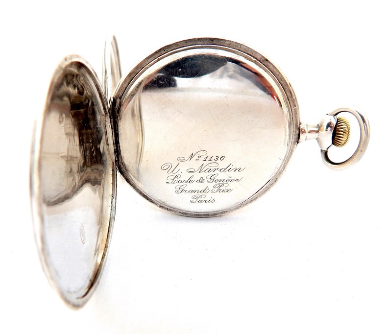 May include: Open silver pocket watch with the back cover ajar, revealing the watch face. The watch has a silver case and a gold-colored winding crown. The watch face has the text "No 1136 U. Nardin, Socle & Gen&egrave;ve, Grands Prix, Paris."