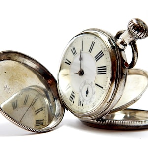 Könnte beinhalten: Eine silberfarbene Taschenuhr mit geöffnetem Klappdeckel, die ein weißes Zifferblatt mit schwarzen römischen Ziffern, ein kleines Hilfszifferblatt für Sekunden und eine geriffelte Aufzugskrone an der Oberseite aufweist.