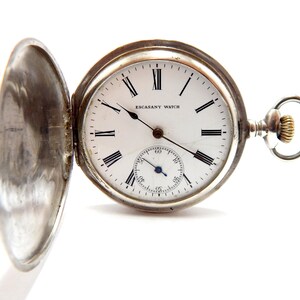 May include: An open silver pocket watch with a white face and black Roman numerals. The watch has a small seconds dial and the words "ESCASANY WATCH" are visible. The watch is set against a white background.