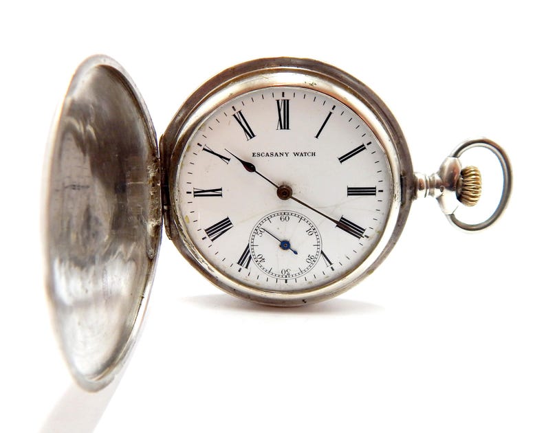 May include: An open silver pocket watch with a white face and black Roman numerals. The watch has a small seconds dial and the words "ESCASANY WATCH" are visible. The watch is set against a white background.