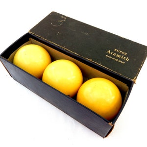 May include: Three yellow billiard balls are presented in an open black box. The box is labelled with the text "SUPER Aramith MADE IN BELGIUM". The balls are a bright yellow colour and are visible inside the box.