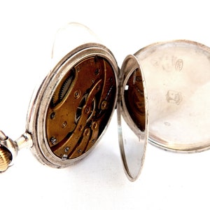 May include: Open silver pocket watch with visible internal gears and a gold-colored winding crown. The watch's cover is open, revealing the intricate mechanics and a shiny silver interior. The watch is set against a white background.