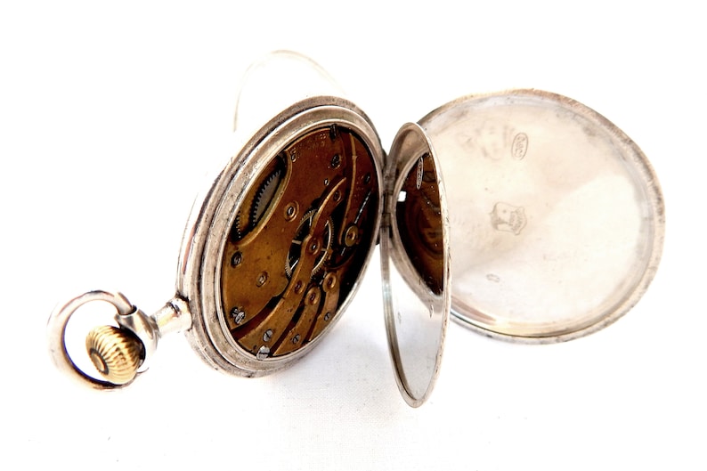 May include: Open silver pocket watch with visible internal gears and a gold-colored winding crown. The watch's cover is open, revealing the intricate mechanics and a shiny silver interior. The watch is set against a white background.