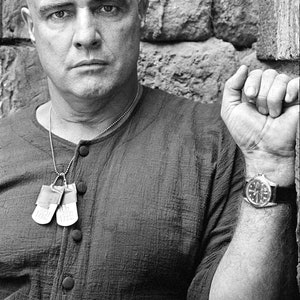 May include: A monochrome portrait of a man with a shaved head, wearing a dark shirt and a wristwatch. He has dog tags and is leaning against a stone wall.