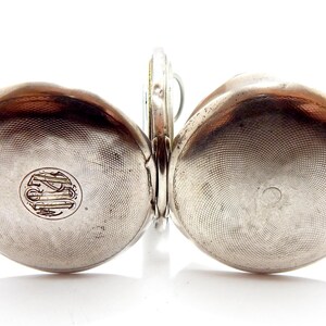 May include: A silver pocket watch with a hinged design, featuring two rounded, textured halves. Each half has a circular emblem. The watch is open, revealing the inner mechanism. The watch is set against a white background.