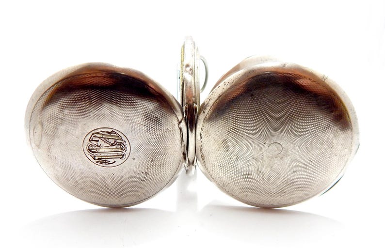 May include: A silver pocket watch with a hinged design, featuring two rounded, textured halves. Each half has a circular emblem. The watch is open, revealing the inner mechanism. The watch is set against a white background.