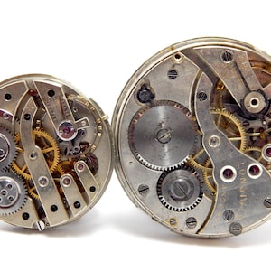 May include: Two antique watch movements with exposed gears and intricate mechanisms. The metal components are silver and gold, with ruby jewels. The watch movements are displayed against a white background.