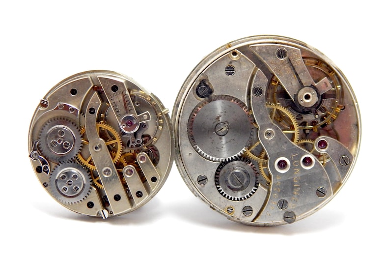 May include: Two antique watch movements with exposed gears and intricate mechanisms. The metal components are silver and gold, with ruby jewels. The watch movements are displayed against a white background.