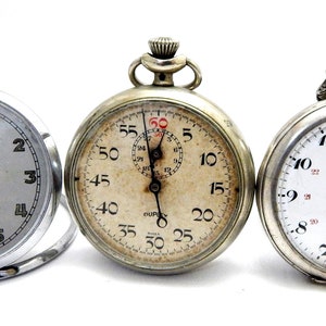 Op de afbeelding: Drie zilveren zakhorloges met witte wijzerplaten. Het horloge links heeft zwarte wijzers en de tekst "Limpia Mágica M. CAS JUANA". Het horloge in het midden is een stopwatch met zwarte wijzers en een kleine wijzerplaat in het midden. Het horloge rechts heeft zwarte wijzers en de tekst "ASTRA" in het midden.
