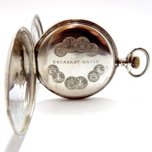 May include: Open silver pocket watch with the back lid ajar, revealing the inner workings. The watch face is engraved with the words "ESCASANY WATCH" and several small circular designs. The watch has a silver case and a gold-colored winding crown.