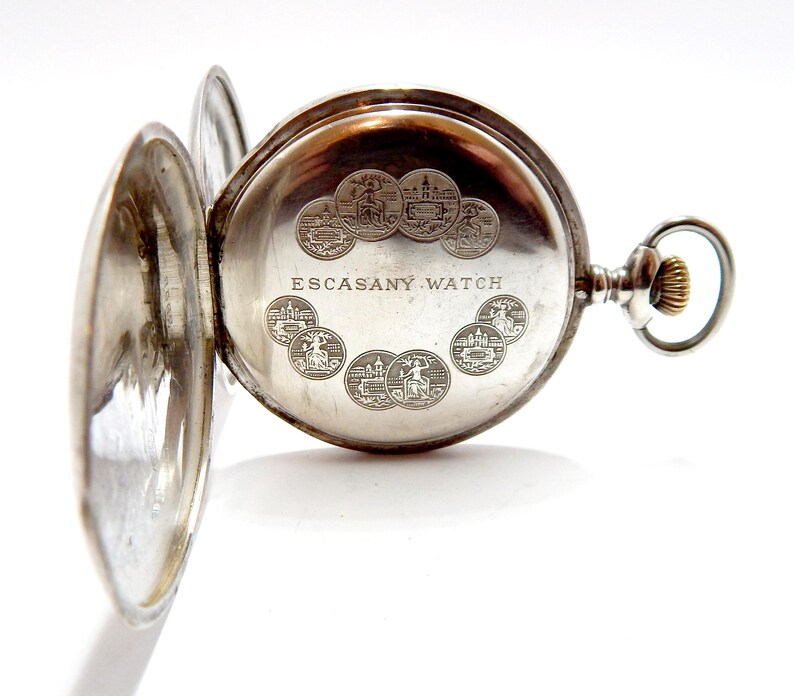 May include: Open silver pocket watch with the back lid ajar, revealing the inner workings. The watch face is engraved with the words "ESCASANY WATCH" and several small circular designs. The watch has a silver case and a gold-colored winding crown.