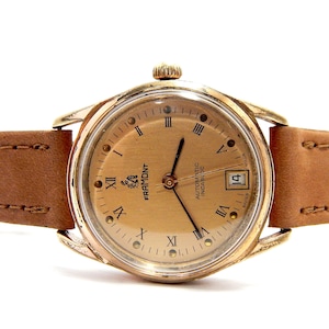 Puede incluir: Un reloj de pulsera dorado con una correa de cuero marrón claro. La esfera del reloj tiene números romanos y una pantalla de fecha. El reloj está marcado con "FRAMONT" y "AUTOMATIC INCABLOC".