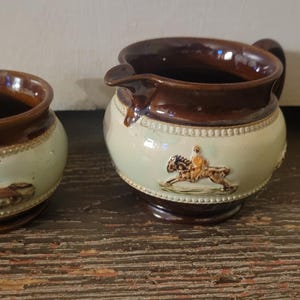 May include: Two antique ceramic pieces: a small bowl and a creamer. Both have a brown rim and base, with a light green body. The creamer features a handle and spout. Each piece has a decorative relief of a horse and rider.