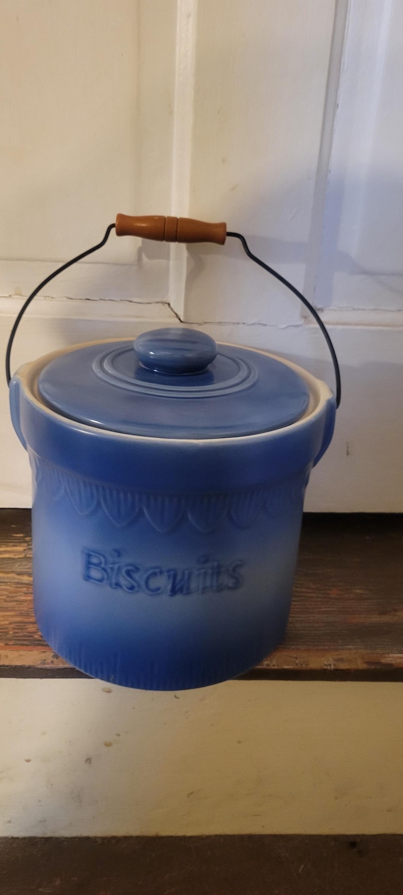 Peut inclure: Pot &agrave; biscuits en c&eacute;ramique bleue avec couvercle et anse m&eacute;tallique avec poign&eacute;e en bois. Le mot "Biscuits" est en relief sur le devant du pot. Le pot mesure environ 18 cm de haut.