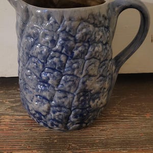 May include: A blue and white ceramic pitcher with a textured, raised pattern. The pitcher has a handle and a spout for pouring. The interior is a darker brown color. The pitcher is sitting on a wooden surface.