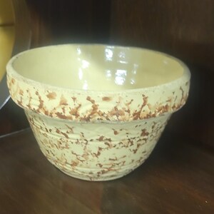 May include: A vintage, cream-colored ceramic mixing bowl with a wide rim and a speckled brown pattern. The bowl has a rounded shape and sits on a dark wooden surface. This kitchenware item is likely used for baking or food preparation.