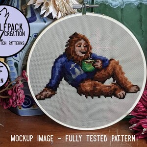 May include: Cross-stitch pattern featuring a relaxed Bigfoot enjoying a mug of coffee. The design is stitched on a light gray fabric, set in a wooden embroidery hoop. The image includes the text "Wolfpack Creation Cross Stitch Patterns" and "PDF Pattern".