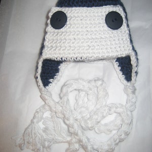 May include: A white and navy blue crocheted hat with two black buttons. The hat has a long white crocheted tassel.