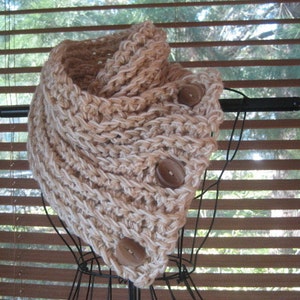 May include: A crocheted cowl in a beige color with three wooden buttons. The cowl is displayed on a black wire stand.