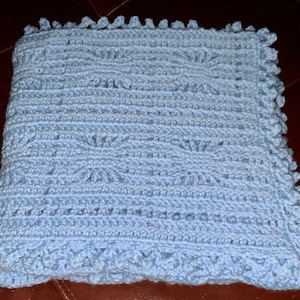 May include: A light blue crocheted blanket with a textured pattern and scalloped edges. The blanket is made of soft yarn and is suitable for a baby or as a decorative throw. The handmade blanket is a cozy and comforting item.