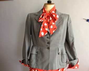 May include: A gray blazer with a red and white floral bow tie. The blazer has a fitted waist and two pockets on the front. The blazer is on a mannequin.