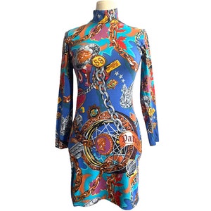 Peut inclure: Une robe bleue à manches longues avec un col montant. La robe présente un imprimé coloré avec des chaînes dorées, des médaillons et le mot "Patent". La robe est présentée sur un mannequin.