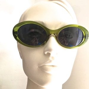 May include: Green oval-shaped sunglasses with dark lenses. The sunglasses are on a white mannequin head.