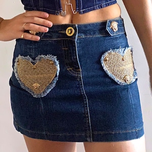 May include: A denim mini skirt with two gold heart-shaped patches. The skirt has a high waist and a zip fastening at the front.