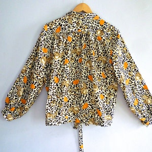 May include: A long-sleeved blouse with a white background and a leopard print pattern in brown and orange. The blouse has a tie at the waist and a button-up collar.