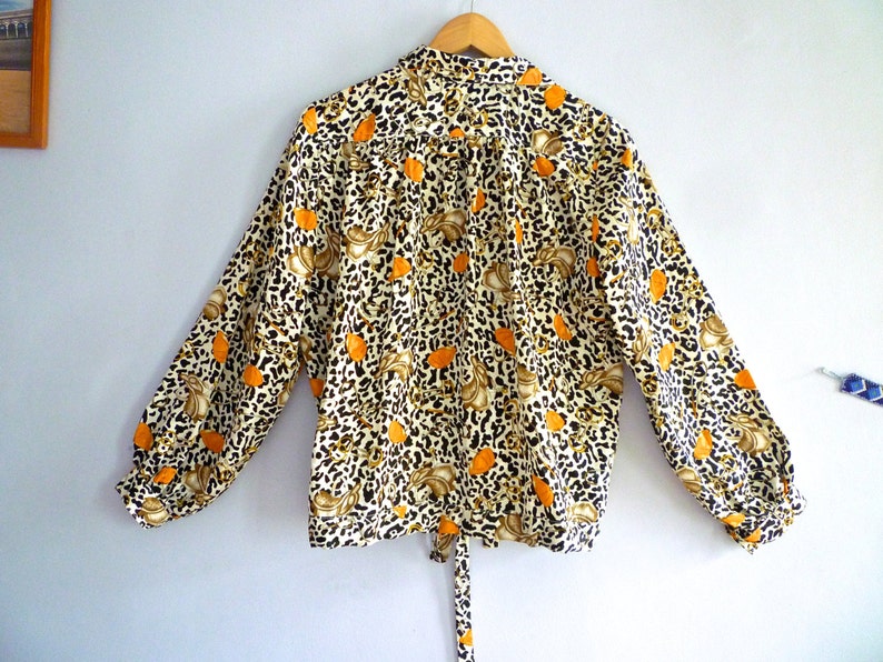 May include: A long-sleeved blouse with a white background and a leopard print pattern in brown and orange. The blouse has a tie at the waist and a button-up collar.