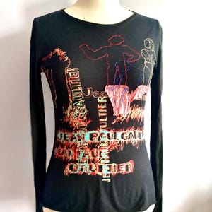 May include: Black long-sleeved shirt with a colourful graphic print of figures and the text "Jean Paul Gaultier".
