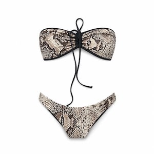 Puede incluir: Un conjunto de bikini de dos piezas con estampado de piel de serpiente en tonos marrón, beige y negro. La parte superior tipo bandeau incluye un detalle de cordón central negro, y la braguita a juego tiene un diseño de pierna alta con ribete negro.
