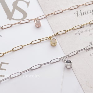 Op de afbeelding: Twee delicate gouden kettingen met kleine ronde hangers met heldere en roze stenen. De kettingen liggen op een wit oppervlak met tekst op de achtergrond.