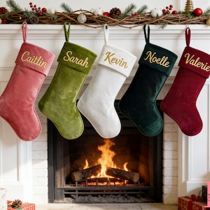 May include: Five velvet Christmas stockings hanging in a row, each featuring a different colour: dusty rose, olive green, white, dark forest green, and deep burgundy. Each stocking is embroidered with a name in gold script: Caitlin, Sarah, Kevin, Noelle, and Valerie.