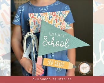 May include: A child holds a blue and green pennant that says "First Day of School" and a yellow pencil that says "3rd Grade". The child is wearing a blue shirt with colorful letters printed on it.