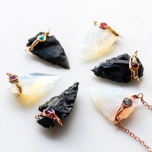 May include: Six arrowhead pendants with gold wire wrapped around them and a small gemstone in the center. The gemstones are turquoise, pink, red, and purple. One of the pendants is on a copper chain.