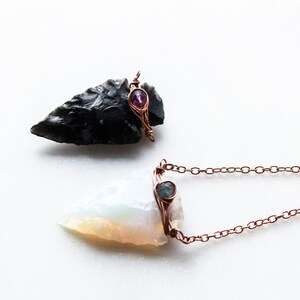 May include: Two arrowhead pendants with copper wire wrapping and gemstone accents. The top pendant is black with a pink gemstone, and the bottom pendant is white with a blue gemstone. The bottom pendant is attached to a copper chain.
