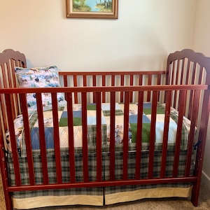 Könnte beinhalten: Ein Gitterbett aus dunklem Holz mit vertikalen Stäben und gewölbten Kopf- und Fußteilen. Das Bett ist mit einer Patchwork-Decke in Grün-, Blau- und Cremetönen, einem passenden Kissen mit Tiermotiv und einem karierten Bettrüschen ausgestattet.