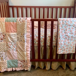 Könnte beinhalten: Ein Kinderbett-Set bestehend aus einer Patchwork-Decke mit rosa, salbeigrünen und floralen Stoffquadraten, teilweise mit strukturierter Noppenoberfläche. Das Set enthält ein passendes florales Spannbettlaken und einen beigen Bettrüschen-Volant.