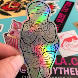 Puede incluir: Una pegatina hologr&aacute;fica de una figura femenina con intrincados dise&ntilde;os de l&iacute;neas. La figura est&aacute; de pie con los brazos a los lados y tiene un dise&ntilde;o en espiral en el centro de su torso. La pegatina es iridiscente y tiene un brillo arco&iacute;ris.