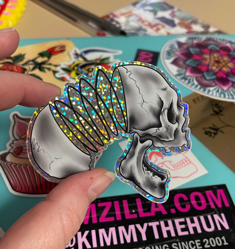 Fingers holding a sticker by the edges - the art on the sticker is a side profile view of a skull that is split in two pieces and glitter fills the void and surrounds the edges of the design