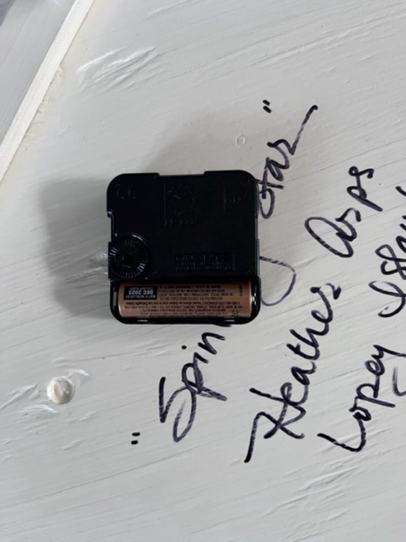 May include: A black plastic quartz clock movement mechanism with a single AA battery inserted into the compartment. The square housing features a central rotating dial for setting the time and is positioned against a white surface with handwritten text.