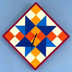May include: A diamond-shaped wall clock featuring a geometric star pattern in shades of orange, yellow, cream, and two tones of blue. The clock has a thin dark red border and two simple black hands positioned at the centre of the star design.
