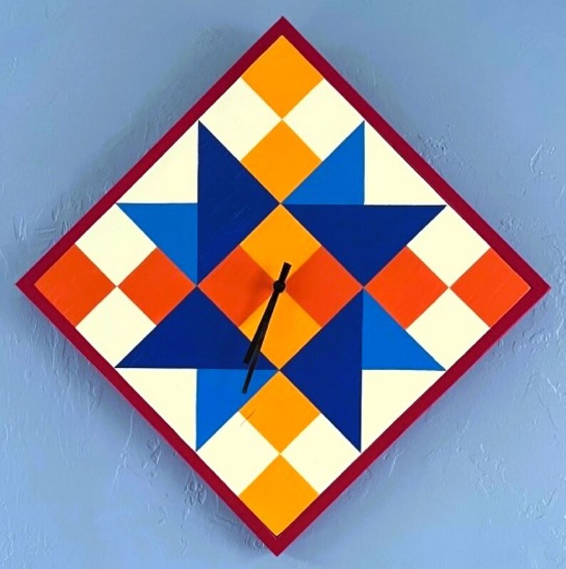 May include: A diamond-shaped wall clock featuring a geometric star pattern in shades of orange, yellow, cream, and two tones of blue. The clock has a thin dark red border and two simple black hands positioned at the centre of the star design.