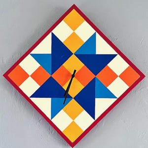 May include: A diamond-shaped wall clock featuring a geometric star pattern in shades of blue, orange, yellow, and cream. The clock has a thin red border and two black hands positioned in the centre. The design is composed of various triangles and squares.