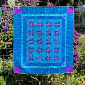 May include: A rectangular quilt featuring a central panel of blue and purple diamond-shaped blocks arranged in a grid. The quilt has a wide blue border with a wavy stitched pattern and solid purple corner squares, all finished with blue binding.
